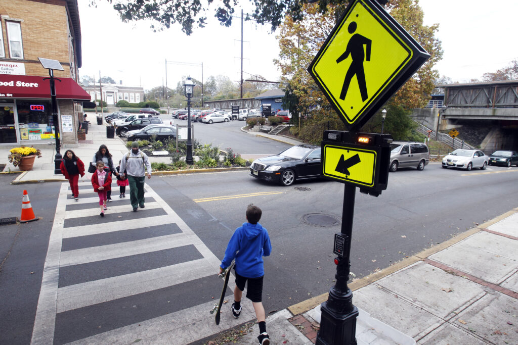 People, including a boy holding a skateboard, walk in a crosswalk while a car stops to wait for them to cross.