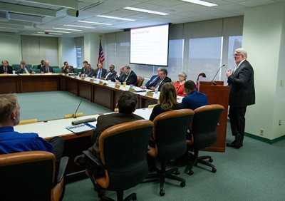 Walter Lane, right, speaks at podium during presentation to Board of Trustees, seated around U-shaped table in front of him.