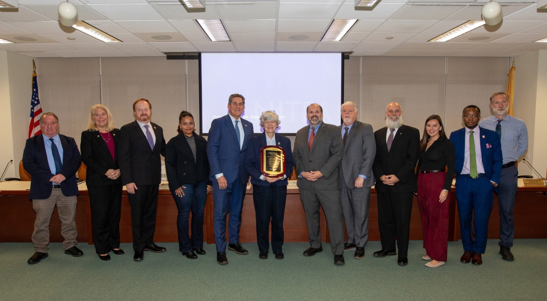 Twelve men and women gather in front of NJTPA Board desk to recognize retiring member holding plaque