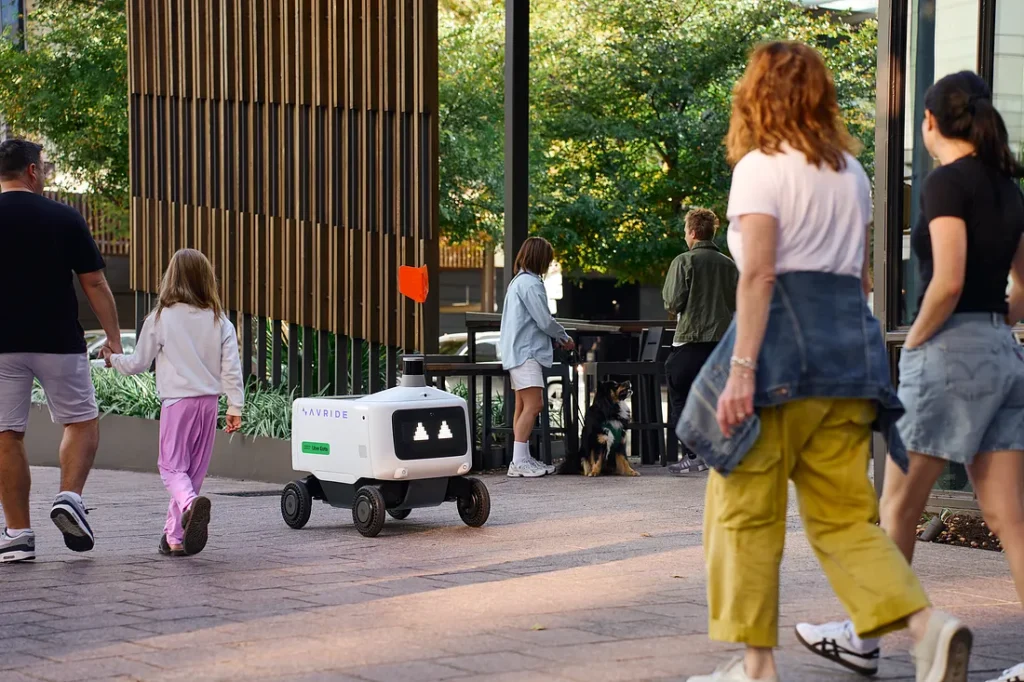 Delivery robot travels on sidewalk amid pedestrians.