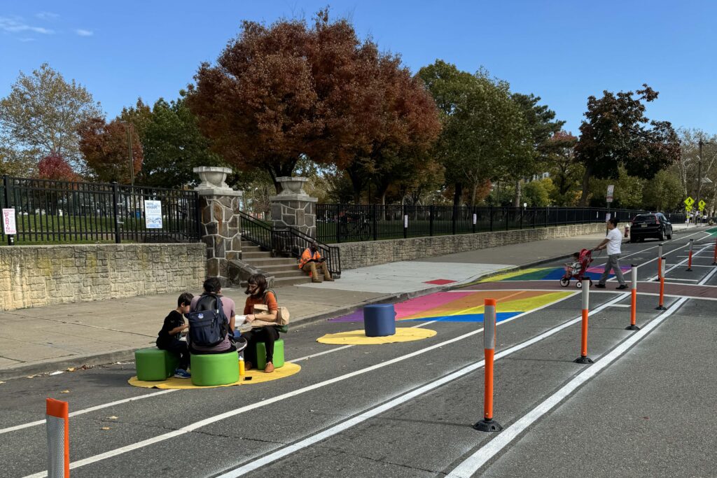 People sit at tables in the street as part of a demonstration project that created temporary bike lanes and additional protected public space to narrow the driving lanes.