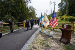 People gather ahead of ribbon cutting ceremony for Highlands Rail Trail with flags and podium in foreground.