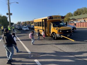 Children board small school bus as traffic pauses behind i and parents look on from crosswalk.