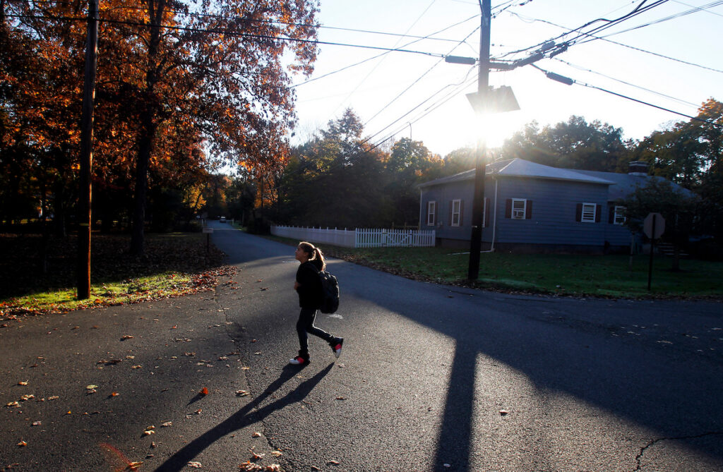 Girl with backpack walks across street at sun rises in background.