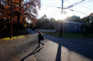 Girl with backpack walks across street at sun rises in background.