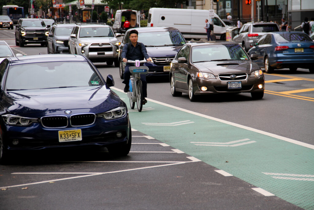 Man rides shared e-bike in bike lane between parked cars and motor vehicle traffic.