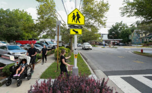 People walking on the sidewalk, pushing stroller, as woman approaches curb to push walk signal button.
