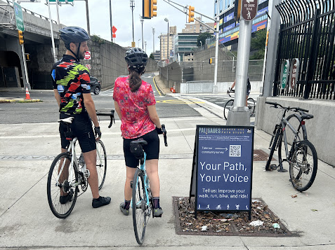 Two people on bikes wait to cross near George Washington Bridge pedestrian, cyclist access near sign about Palisades Shared Use Path STudy.
